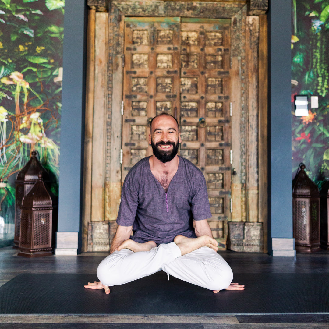 Man sitting in a meditative pose in front of an ornate wooden door with a colorful ceiling.