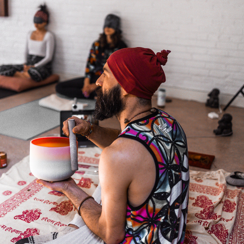 Man with a red turban sitting on a bed in a room with white walls and another person in the background.